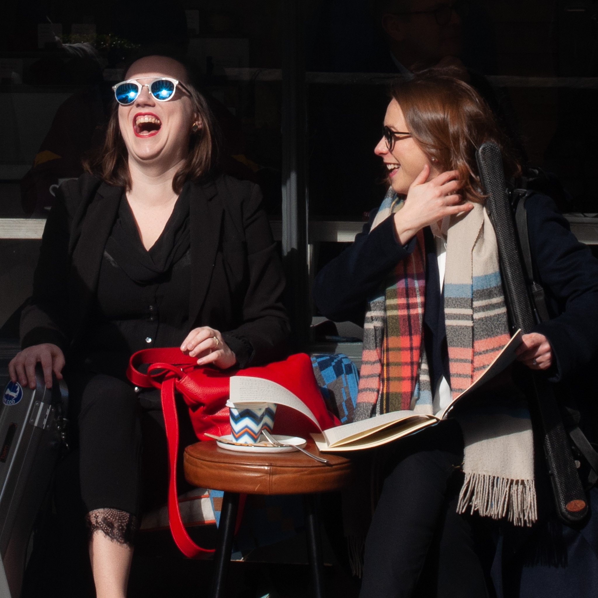 Paula and Rachel laughing. Paula is wearing blue-lensed sunglasses and concert blacks, her left hand resting on a red leather bag with a cup of coffee in front of it. Rachel is turned to face Paula, wearing concert blacks and a dark coat, with a light-coloured scarf with irregular coloured stripes. She is holding an open score.